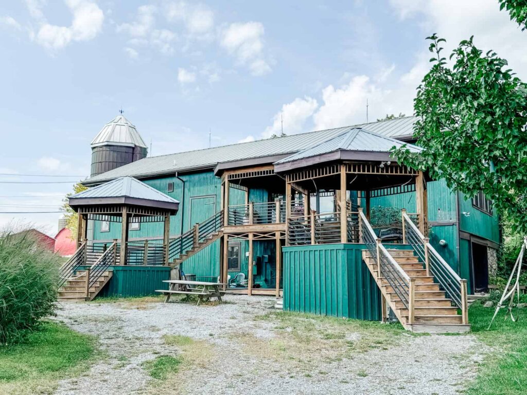 A green-painted wooden building with a unique multi-level deck and a silo, serving as the main structure of the Heron Hill Winery, with a picnic bench and gravel path in the foreground.