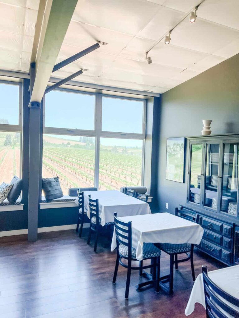 An inviting tasting room with simple white tablecloths on wooden tables, offering panoramic vineyard views through large windows. The room features dark wood flooring and a cozy, modern design, ideal for enjoying wine indoors.