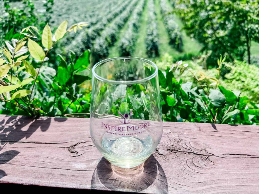 A clear glass featuring the "Inspire Moore" logo, perched on a wooden railing with green foliage and grapevines visible in the background.