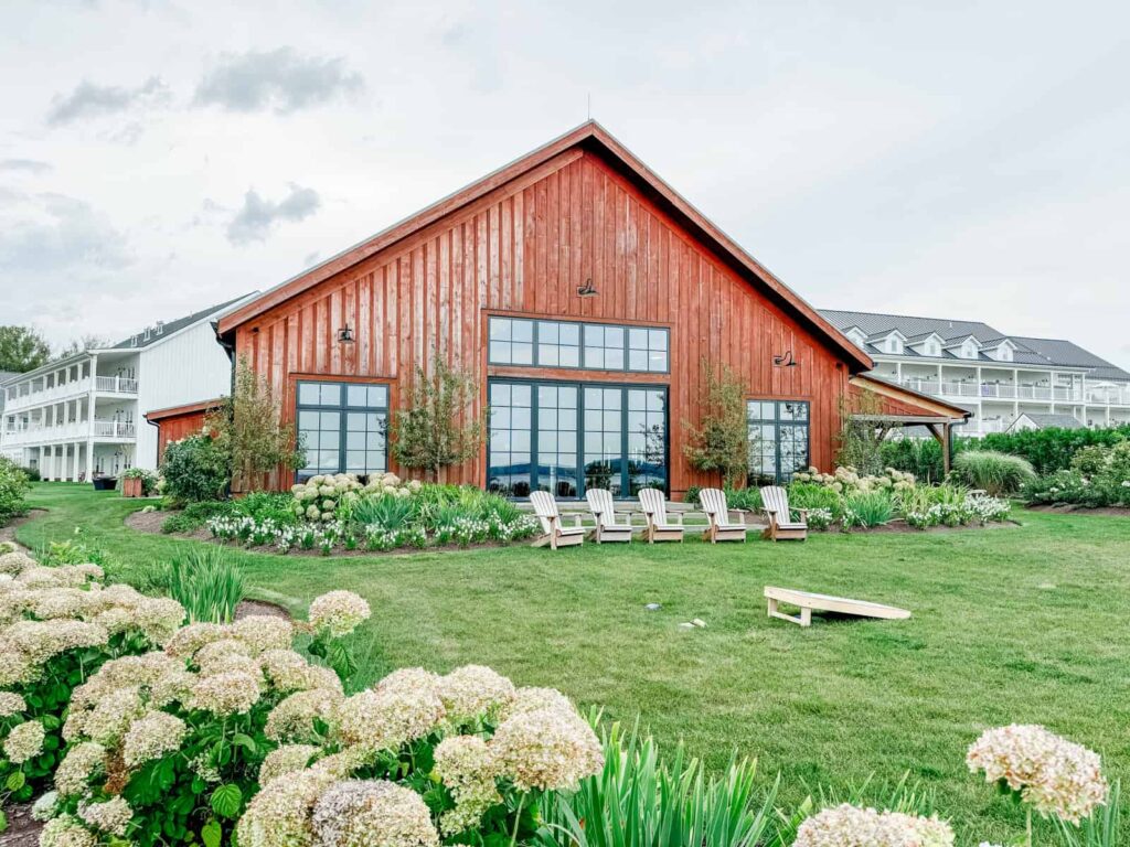 A wooden barn-style building with large glass windows and neatly landscaped gardens. Adirondack chairs are arranged in front of the building, and a cornhole game board is set up on the grass. A white multi-story hotel building is visible in the background.