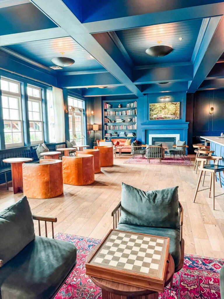 A cozy library room with navy blue walls and ceiling, featuring a fireplace, bookshelves, and modern seating with plush chairs and couches. A chessboard sits on a small table in the foreground, surrounded by vibrant area rugs.