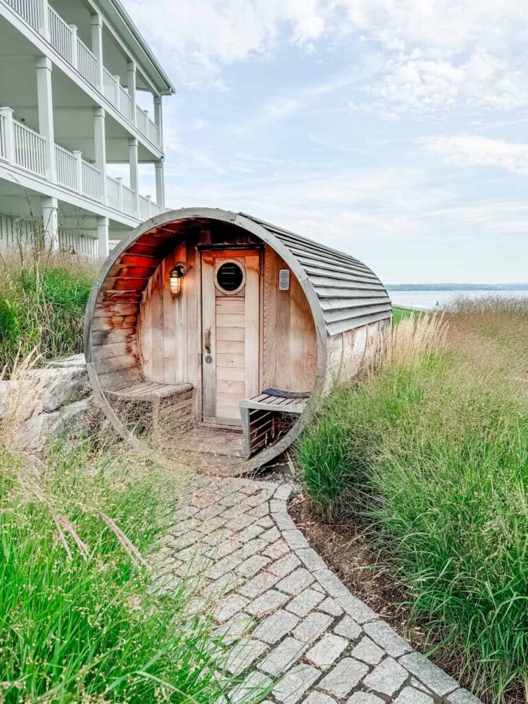 A barrel-shaped wooden sauna nestled among tall grasses and stone paths. A multi-story white hotel building is visible to the left, and the serene lakeside is in the distance.