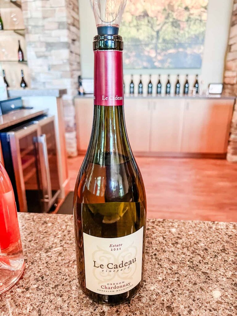 A bottle of Le Cadeau 2021 Chardonnay sits on a granite counter at the winery's tasting room, with a wine aerator attached to the top and other wine bottles in the background.