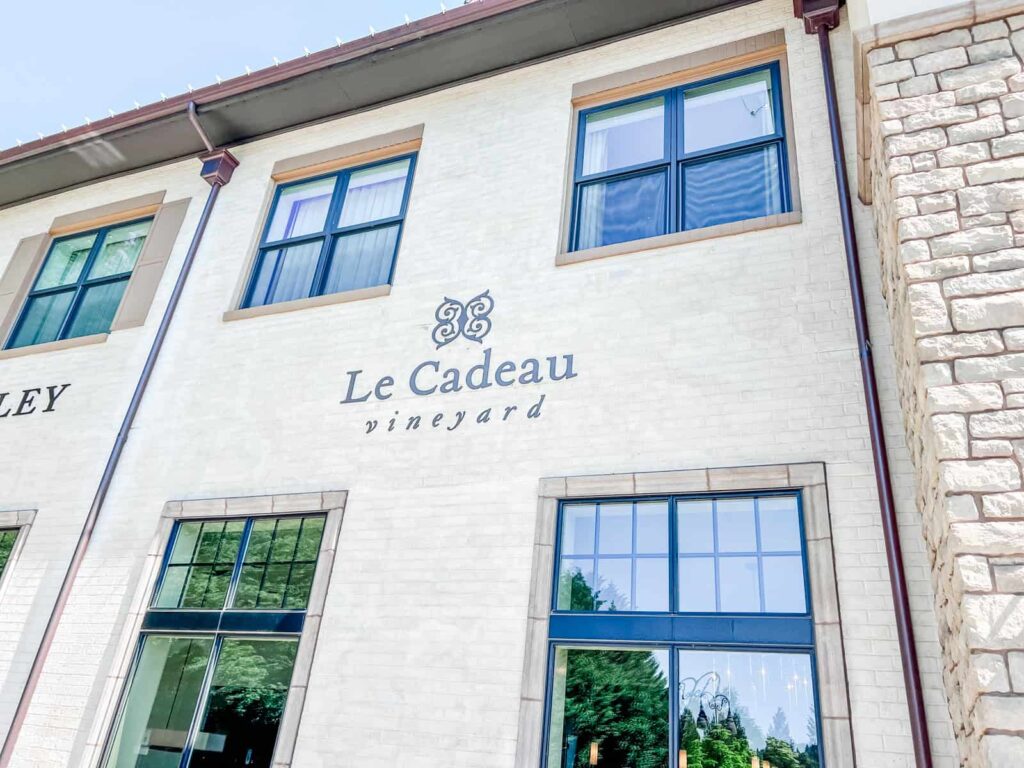 The exterior of Le Cadeau Vineyard with its name prominently displayed on the cream-colored brick building, featuring large windows and elegant lettering.