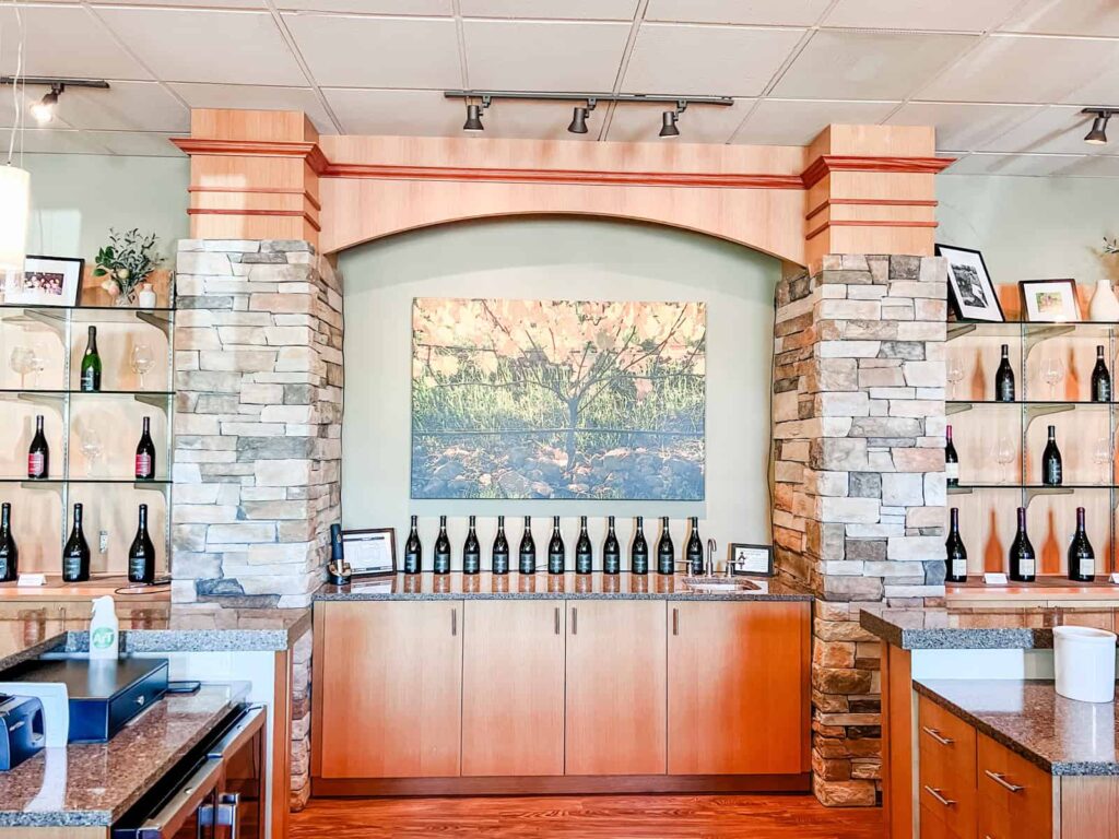 The interior of Le Cadeau's tasting room, with bottles of wine lined up on a wooden counter, flanked by stone pillars and shelves displaying wine bottles and glasses. A large painting of a vineyard scene hangs above.