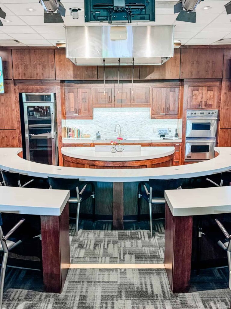 A modern demonstration kitchen setup with a semi-circular countertop, bar seating for guests, a central cooking area, and overhead lighting, creating a classroom-style environment for culinary demonstrations.