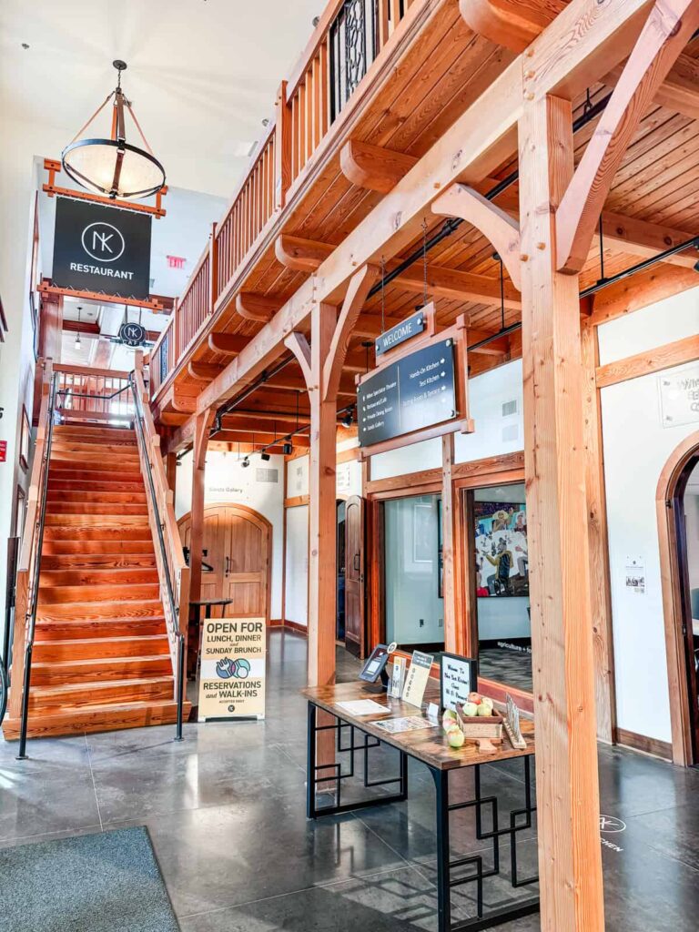 A bright, open lobby with wooden beams and railings, featuring a staircase, a welcoming table with brochures and apples, and signs for NK Restaurant and Wine Spectator Theater.
