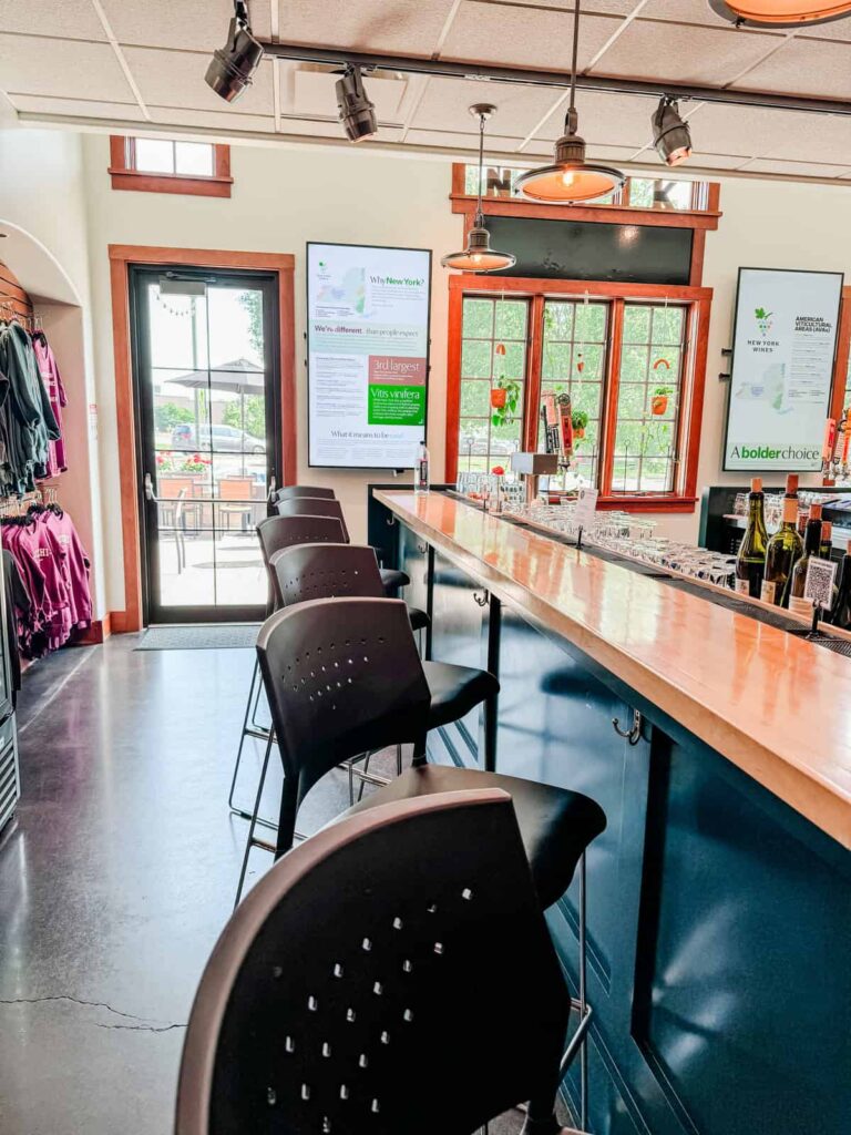 A sleek bar with high chairs and a window displaying New York wine region facts. Bottles of wine and tasting menus are visible along the bar.