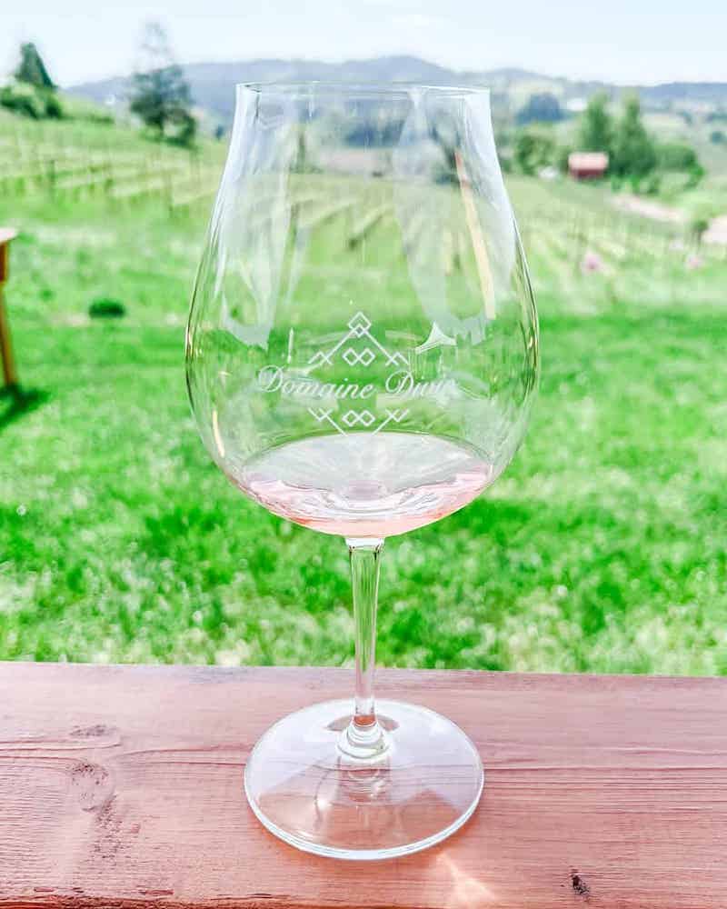 A wine glass with the Domaine Divio logo filled with white wine, set on a wooden table with a Ribbon Ridge vineyard view in the background.