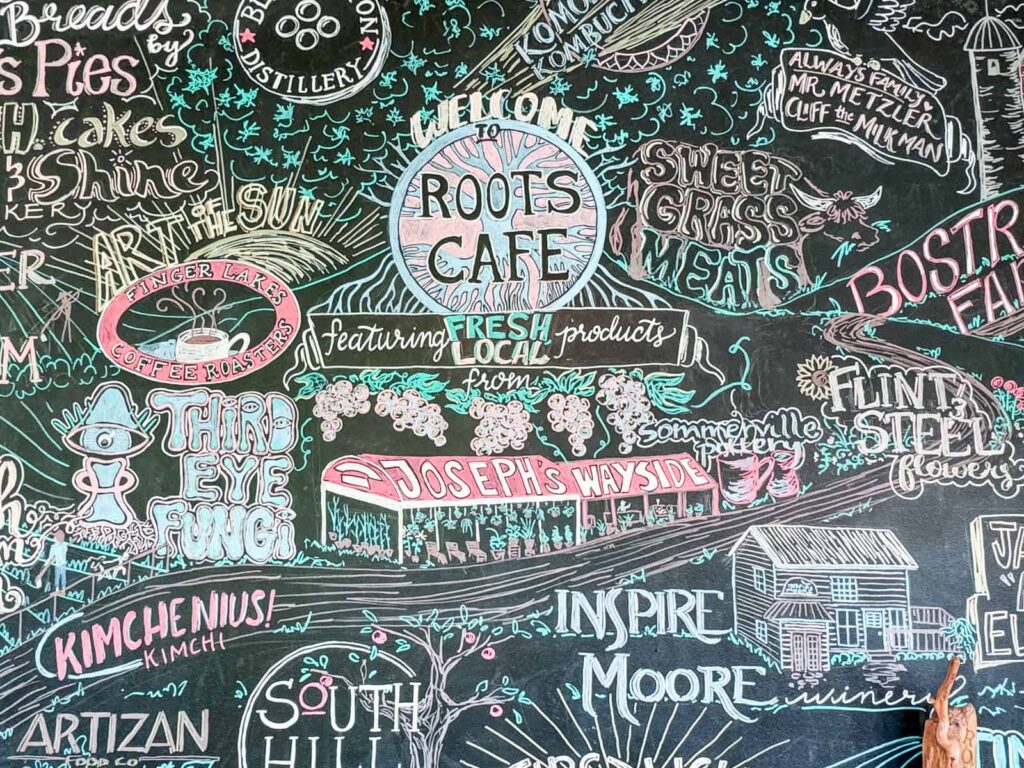 A vibrant chalkboard mural at Roots Café featuring artistic logos and drawings for local businesses, such as Finger Lakes Coffee Roasters, Sweet Grass Meats, and Inspire Moore Winery.