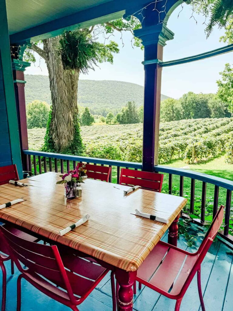A cozy outdoor dining table set for six with red chairs and colorful table settings on a wraparound porch, overlooking a vineyard and a scenic mountain in the background.