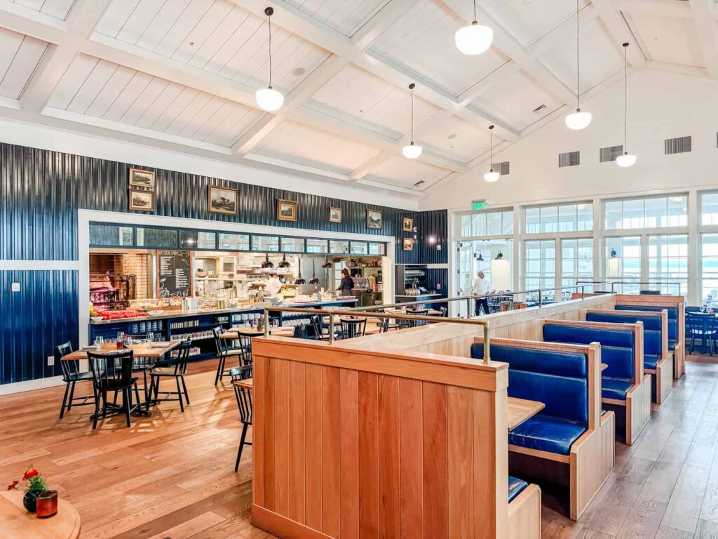 A bright and spacious dining area at Rose Tavern, featuring wooden booths with cushioned seats, wooden tables, and an open kitchen view with a chalkboard menu.