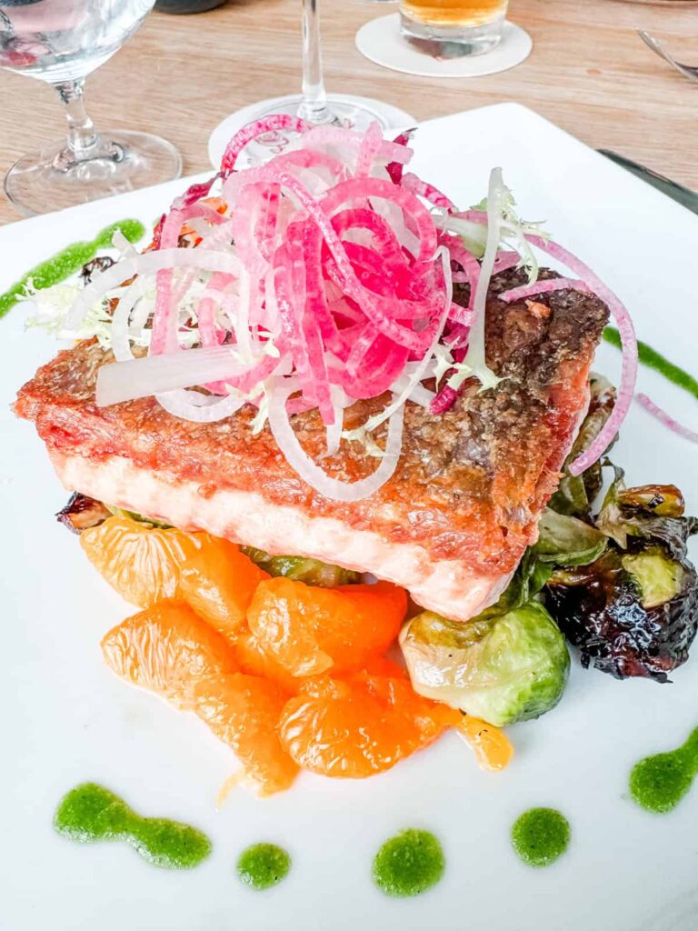 Crispy-skinned salmon served on a bed of roasted Brussels sprouts and vibrant orange segments, garnished with thinly sliced red onions and microgreens.
