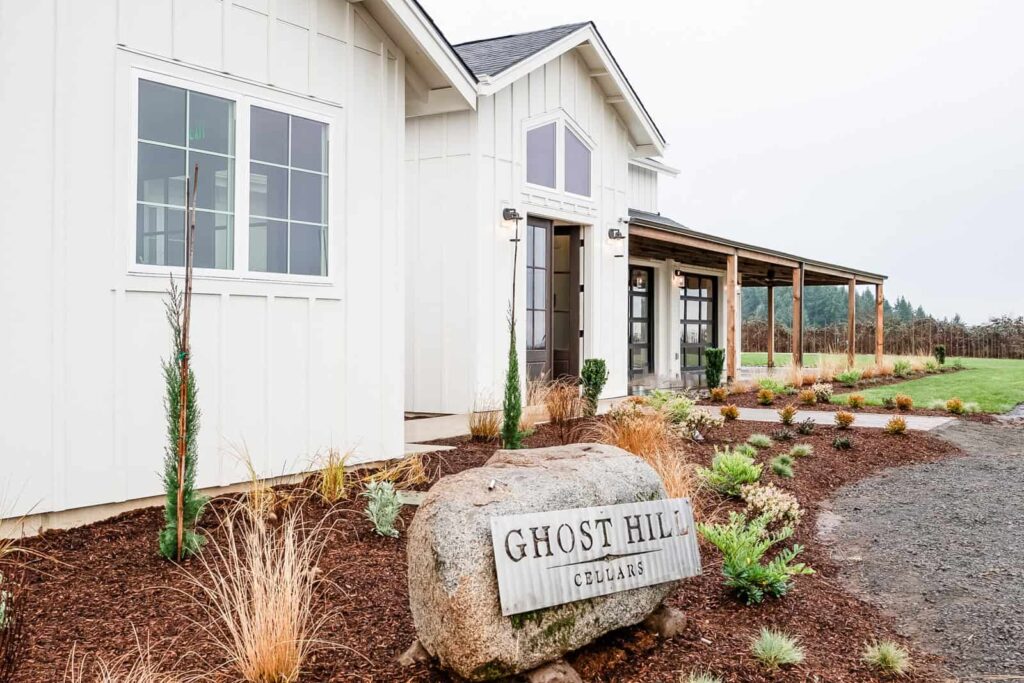 A modern white building with a farmhouse style design, featuring a porch with wooden posts and landscaping of ornamental grasses and shrubs. A large rock with "Ghost Hill Cellars" engraved sits near the entrance.