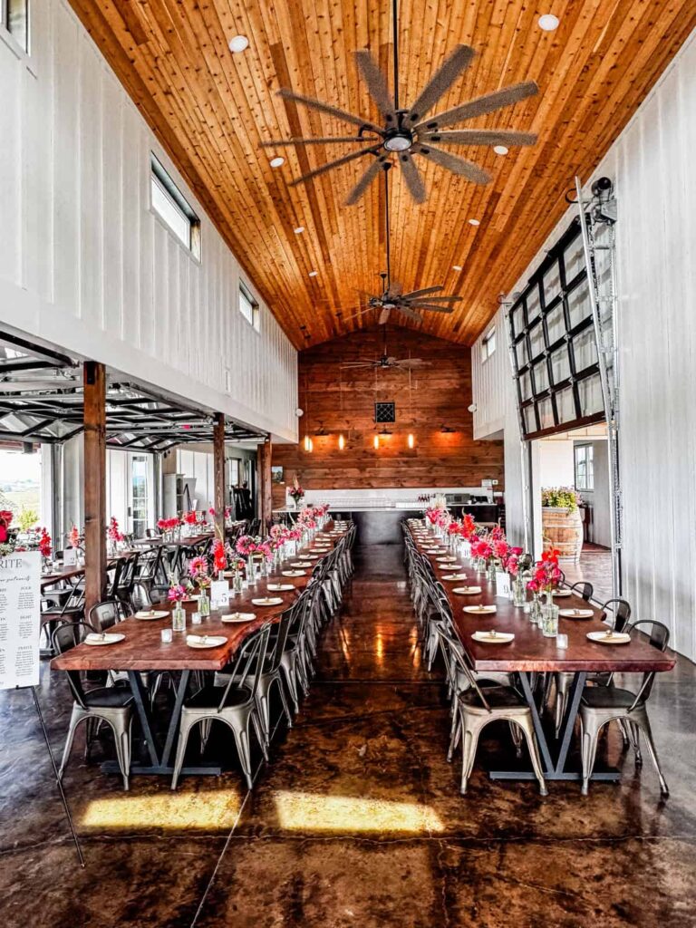 A long, rustic indoor tasting area with polished wooden tables set with plates and vases of flowers. The room features high wooden ceilings and industrial-style fans.