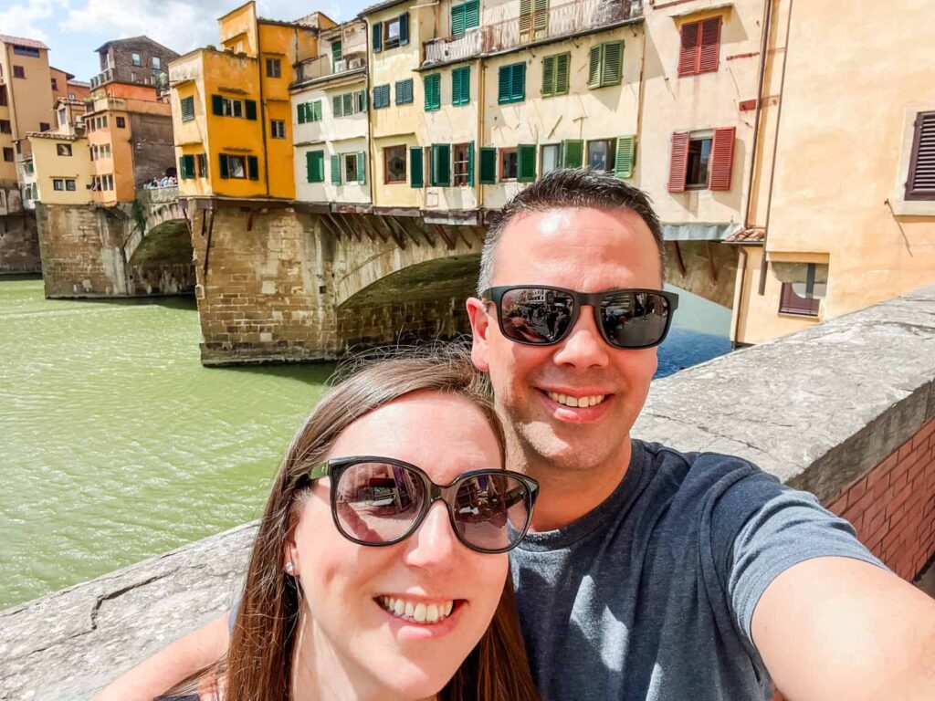 Laura and her husband taking a selfie in front of Ponte Vecchio, Florence's iconic bridge with colorful buildings reflecting off the Arno River.