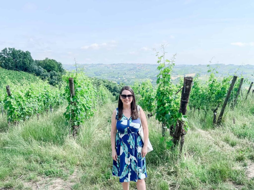 Laura in a blue patterned dress standing among lush, green vineyard rows on a sunny day, overlooking a scenic landscape.