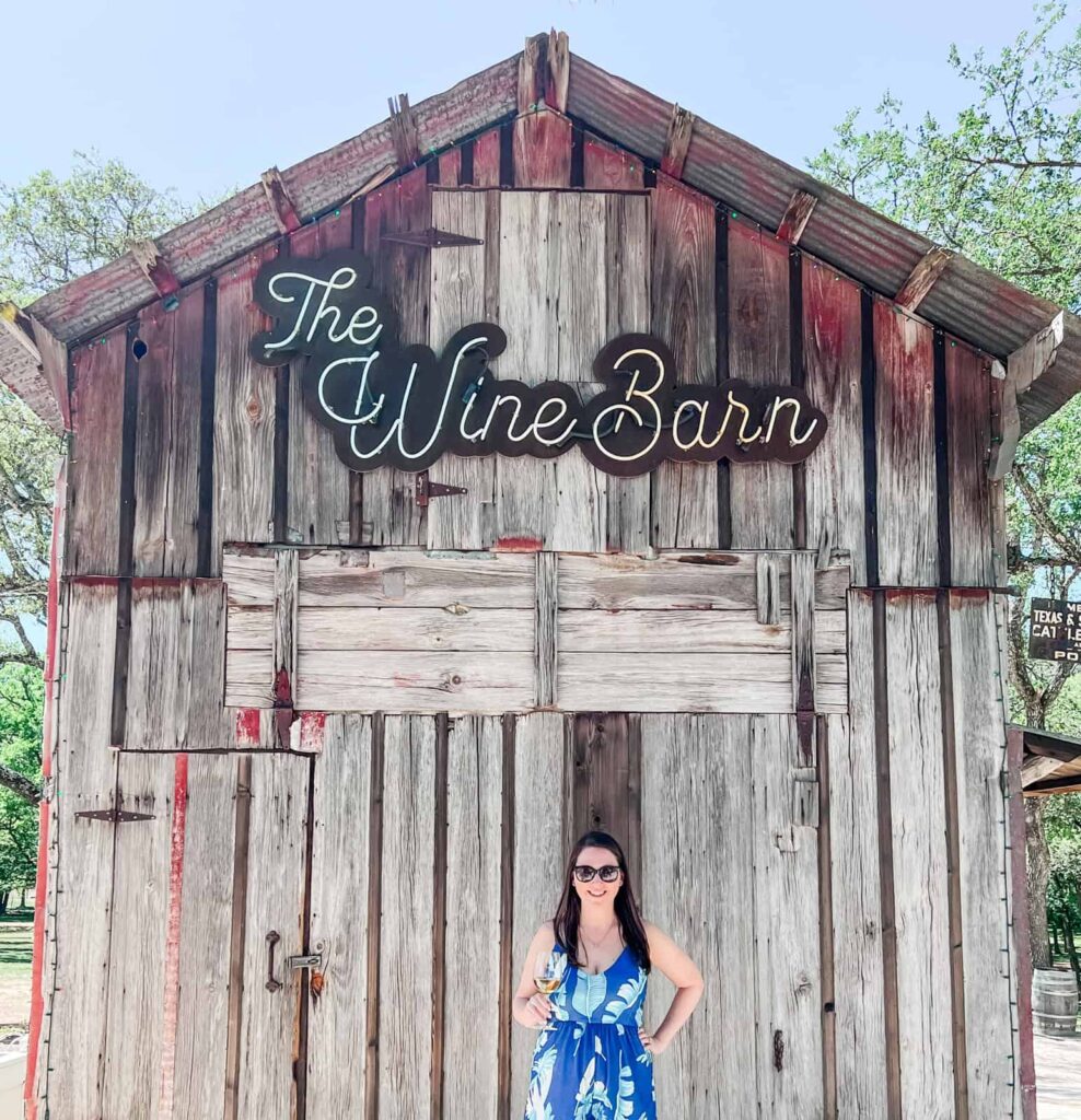 Laura holding a glass of wine in front of a rustic wooden building with a sign reading “The Wine Barn.”