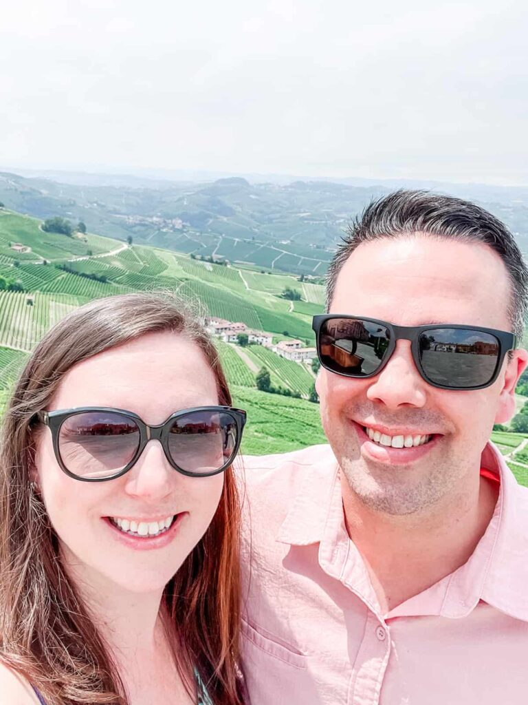 Laura and her husband smiling together with a view of expansive vineyards and hills under a bright sky in the background.