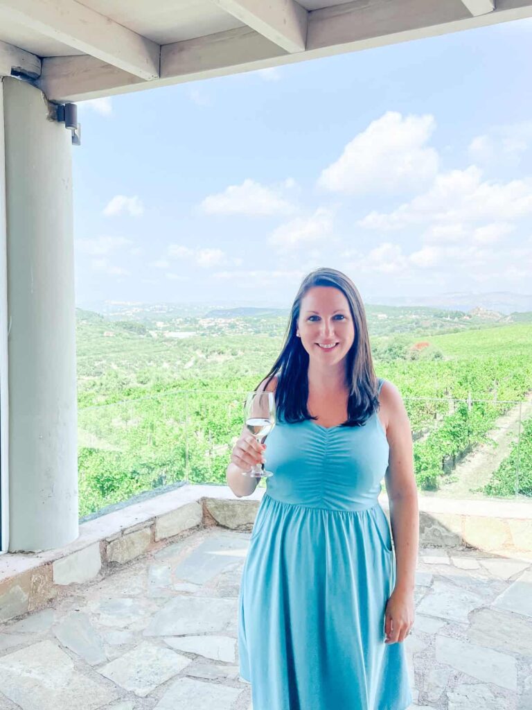 Laura in a blue dress holding a wine glass, standing under a shaded patio with vineyard views stretching out behind her.