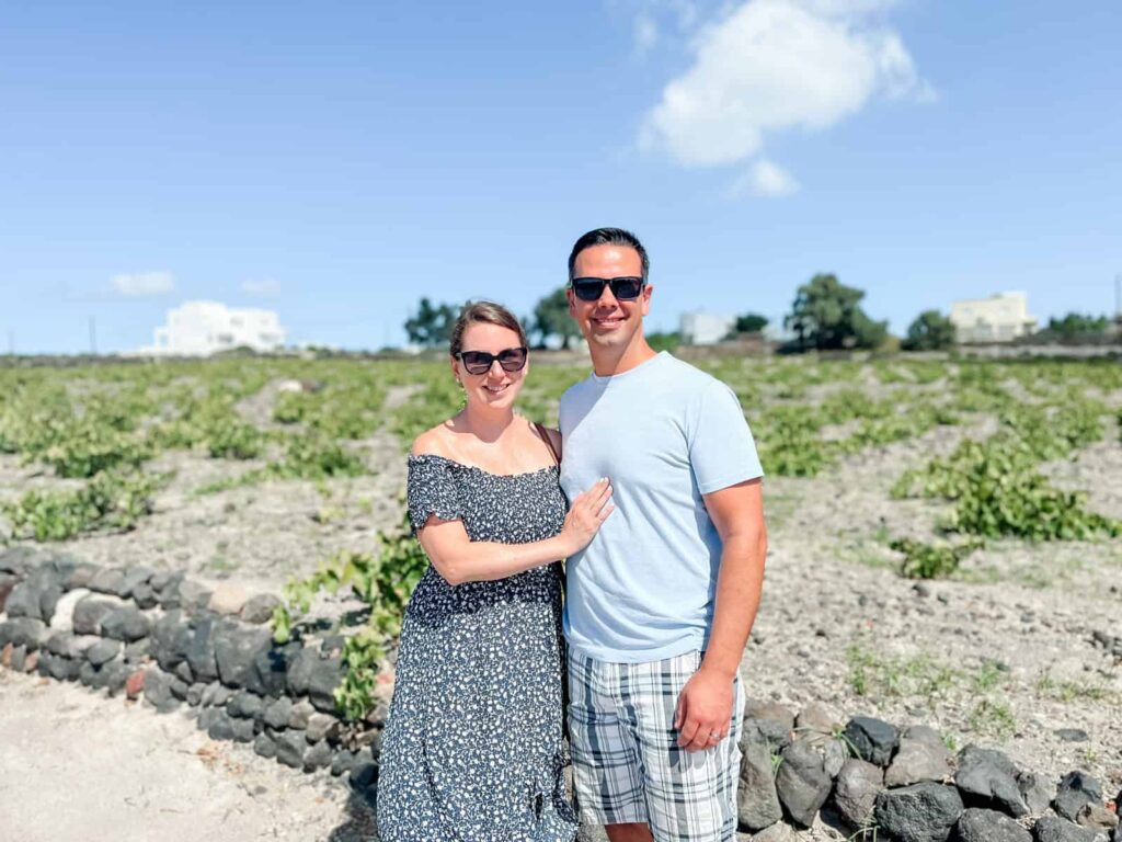 Laura and her husband posing outdoors in a Santorini vineyard landscape under a clear blue sky, and traditional whitewashed buildings in the background.