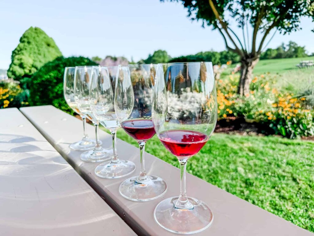 A row of wine glasses, some partially filled with red and white Finger Lakes wines, lined up on a white table with a garden background, featuring vibrant green foliage and blooming flowers.
