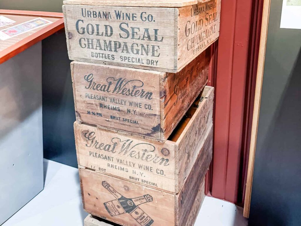 A stack of vintage Finger Lakes wooden wine crates featuring labels from Urbana Wine Co. and Pleasant Valley Wine Co., advertising "Gold Seal Champagne" and "Great Western Brut Special." The aged wood and typography evoke a rustic, historical charm, displayed in a corner of a room.