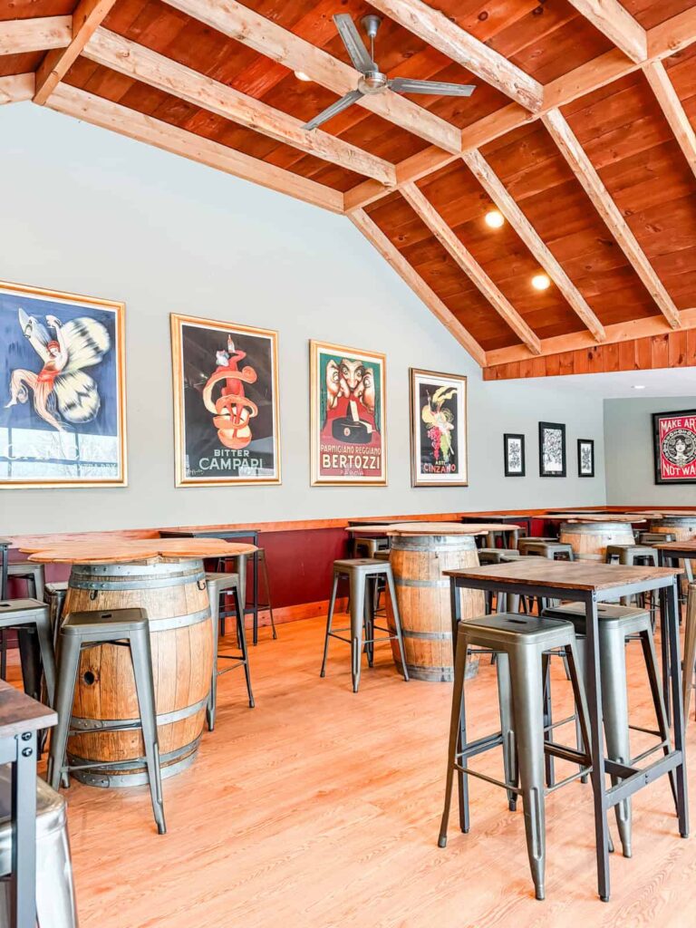 A tasting room with a vaulted wooden ceiling, wine barrel tables, metal stools, and colorful vintage posters decorating the walls.