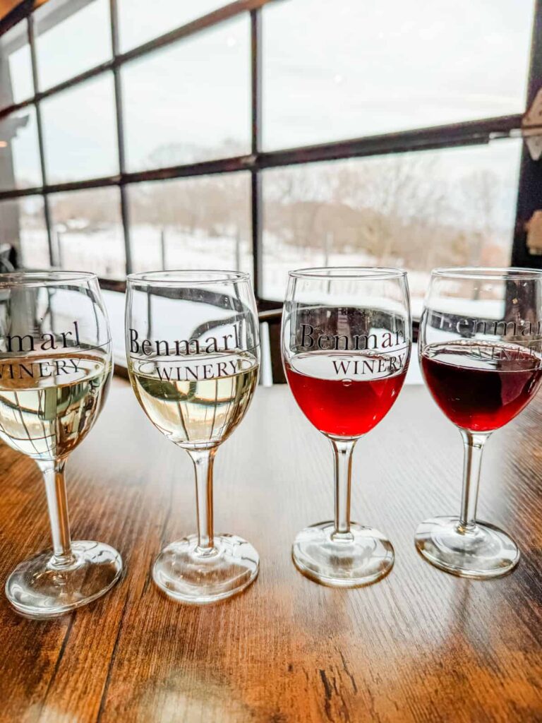 A row of four wine glasses labeled "Benmarl Winery," filled with a progression of white to red wines, placed on a wooden table near a large window overlooking a snowy vineyard.