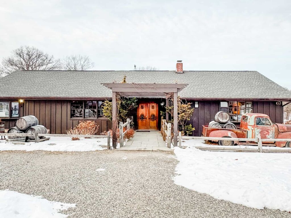 The exterior of the Benmarl Winery tasting room, a rustic brown building with a snow-covered entrance, wooden barrels, and an old red pickup truck in the foreground.