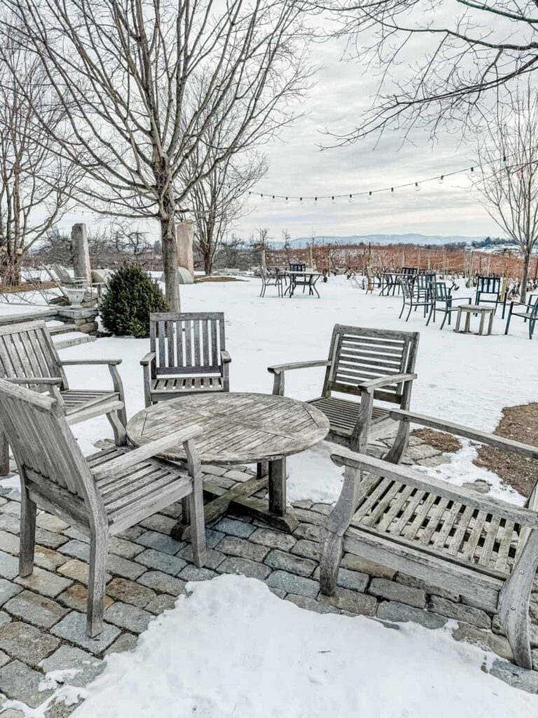 A rustic outdoor seating area covered in snow, featuring weathered wooden chairs and tables. Leafless trees and vineyard rows stretch into the background, with string lights overhead and a cloudy winter sky.