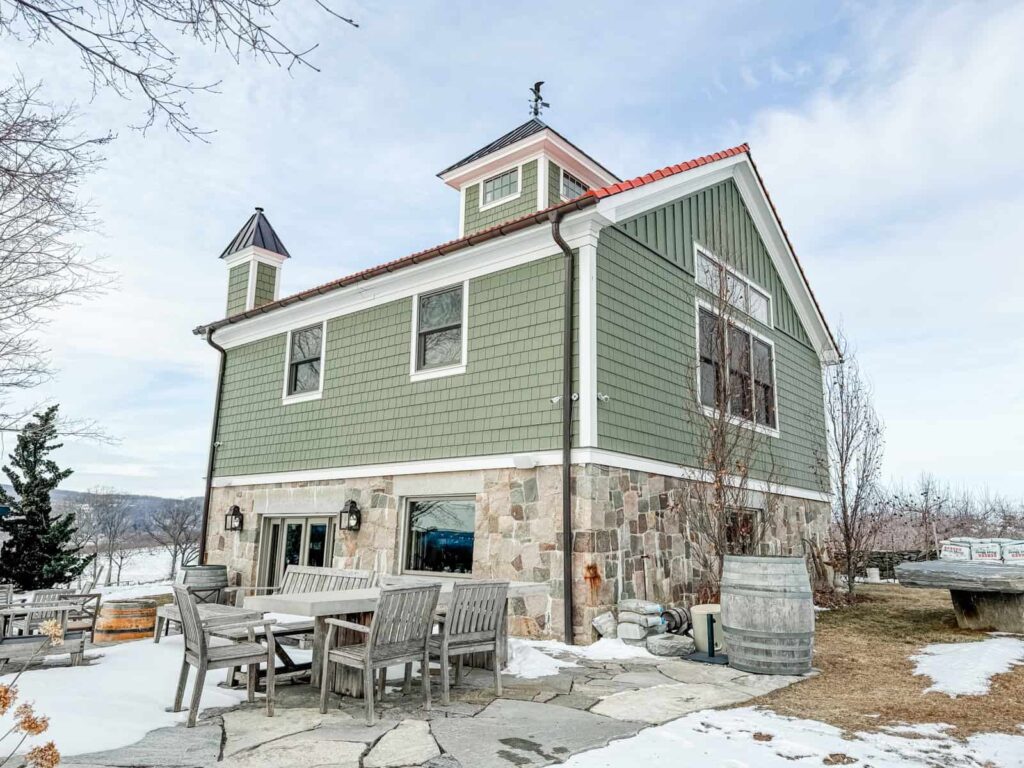 A charming two-story tasting room with green siding and a stone foundation. Outdoor seating, a wooden wine barrel, and patches of snow surround the building, with scenic hills in the distance.
