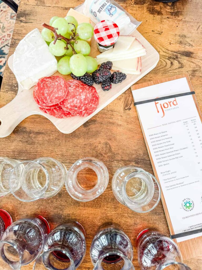 A wooden table set with an elegant charcuterie board featuring green grapes, blackberries, cheese, salami, and a small jar of jam. A tasting flight of red and white wines is arranged in front of a Fjord Vineyards menu.