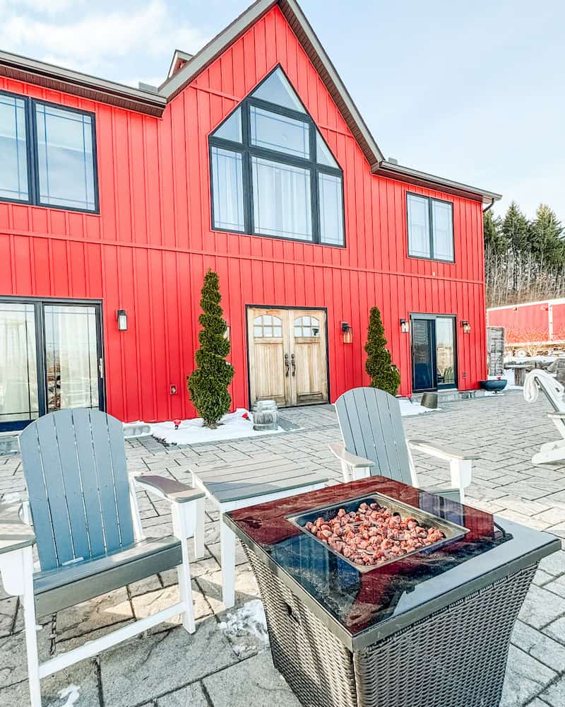 A vibrant red barn-style building at Nostrano Vineyards in the Hudson Valley. In the foreground, two Adirondack chairs sit beside a modern fire pit on a paved patio.