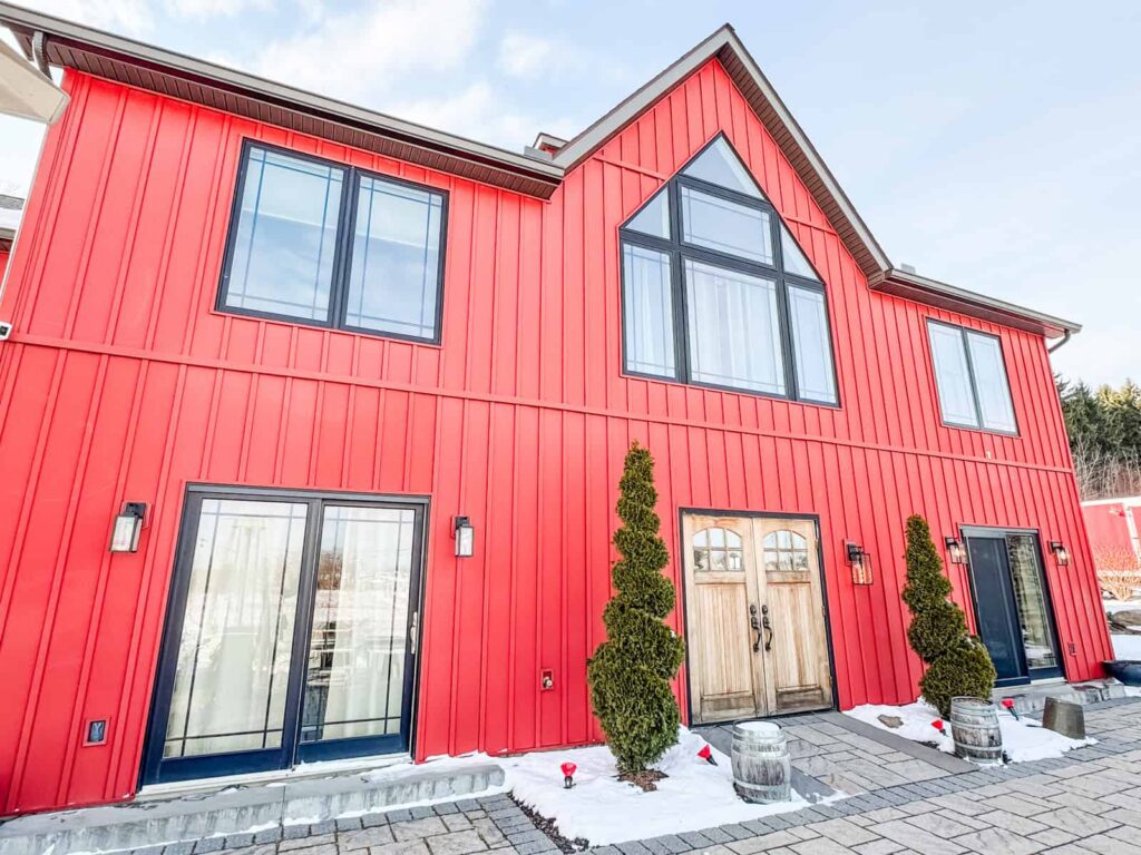 A bright red modern barn-style winery building with black window frames, wooden double doors, and evergreen shrubs lining the entrance.