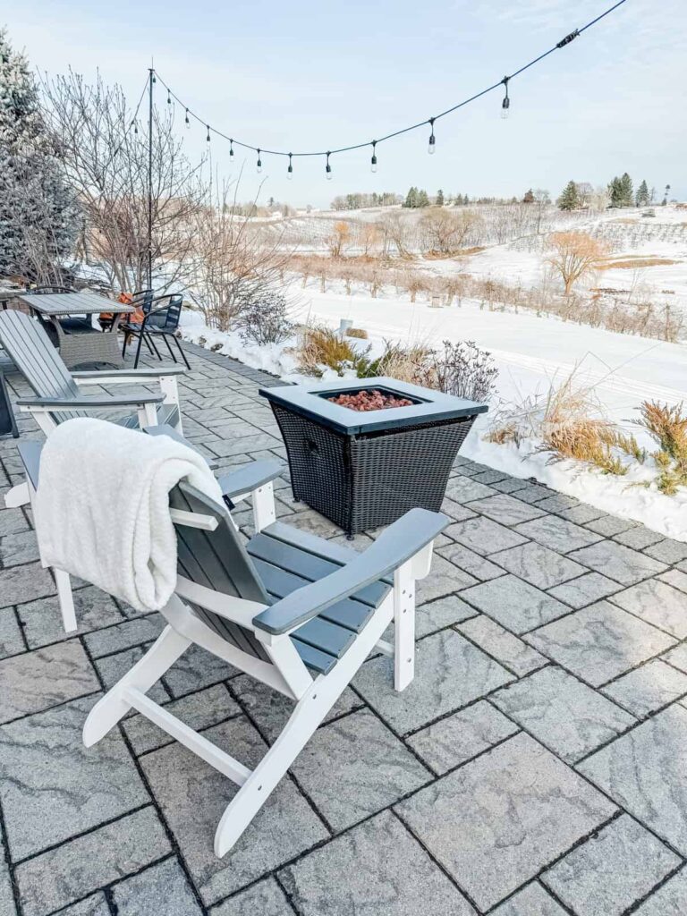 n outdoor winter seating area with Adirondack chairs, a blanket draped over one chair, a fire pit, and string lights hanging above. Snow-covered vineyards are visible in the distance.