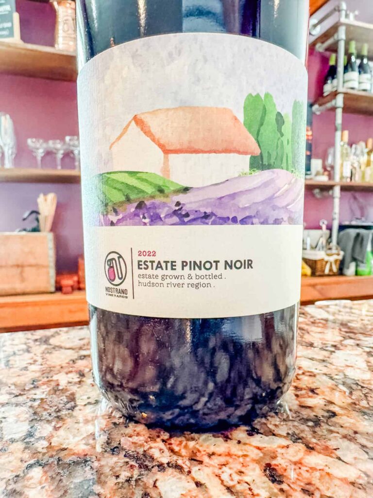 A bottle of Nostrano Vineyards 2022 Estate Pinot Noir, featuring a watercolor-style label depicting a vineyard with a red-roofed house, sitting on a marble countertop.