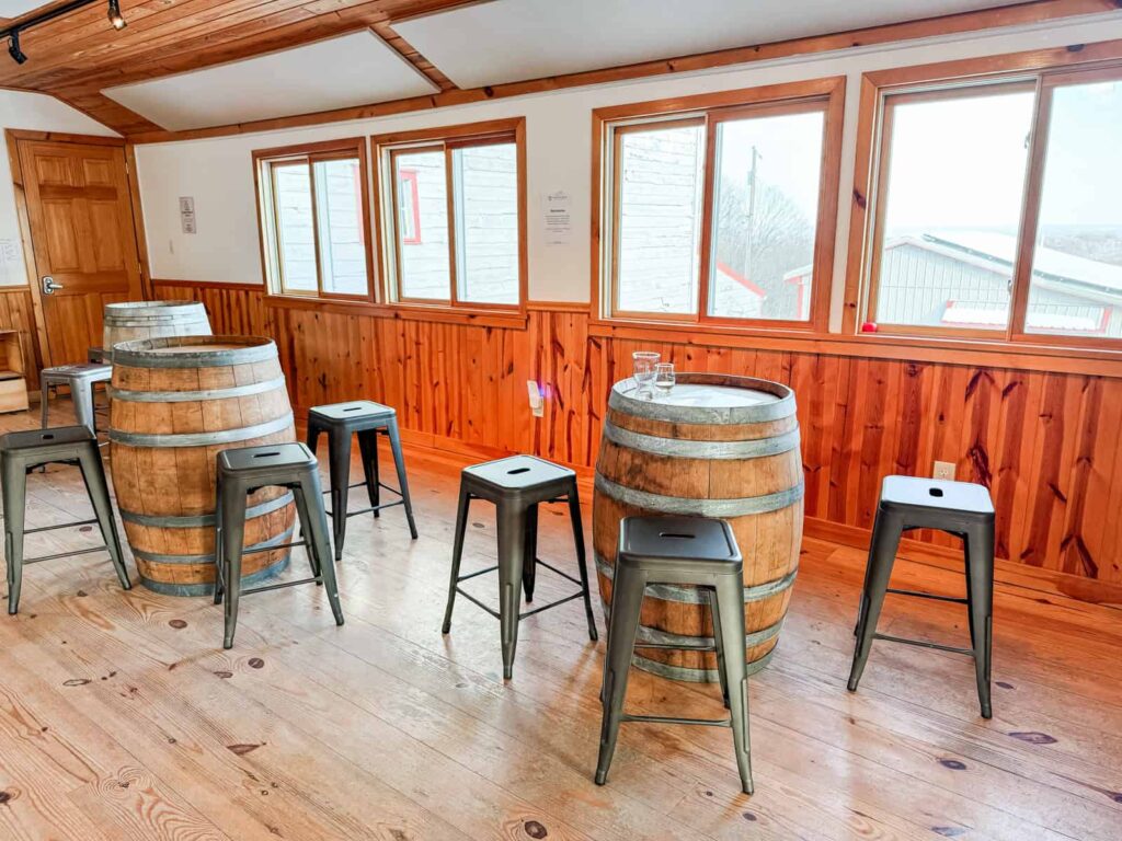 A cozy tasting room at Quartz Rock Vineyard with wooden barrel tables, black metal stools, wood-paneled walls, and large windows overlooking the vineyard.