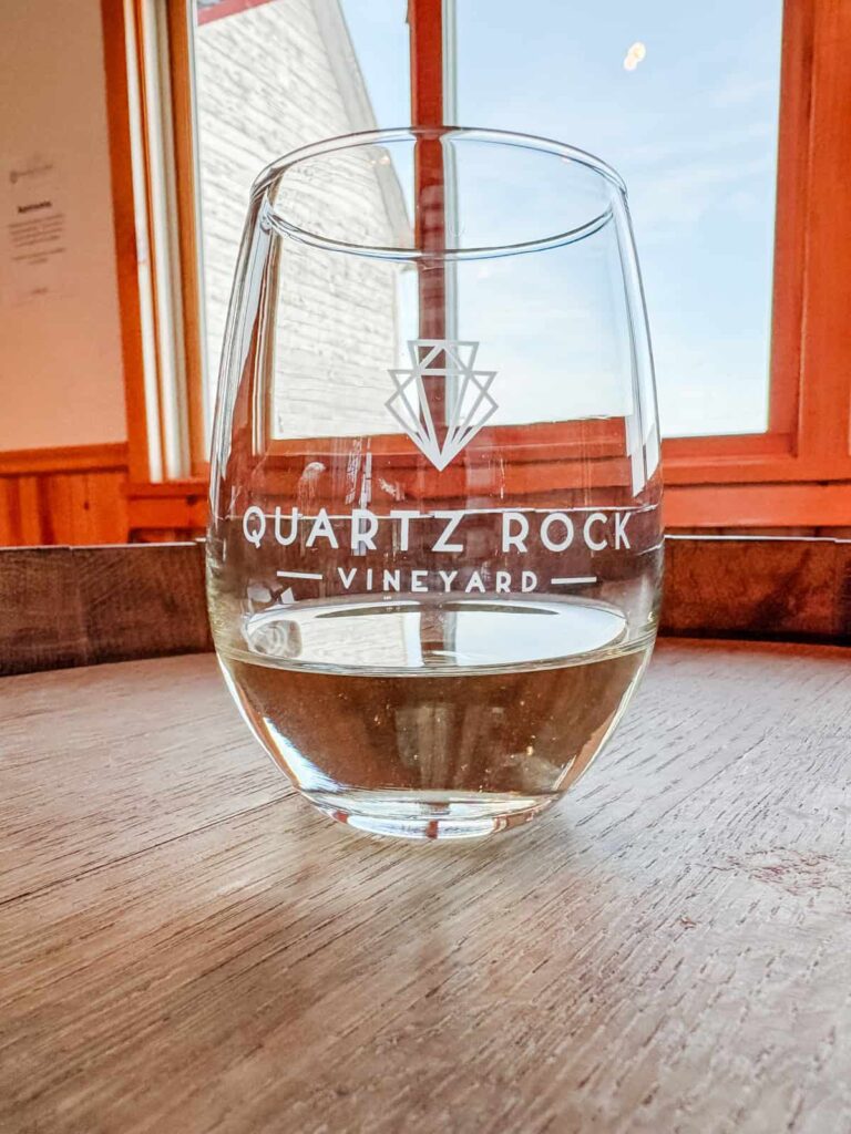 A glass of white wine with the "Quartz Rock Vineyard" logo etched on it, placed on a wooden barrel table with a bright window view behind it.