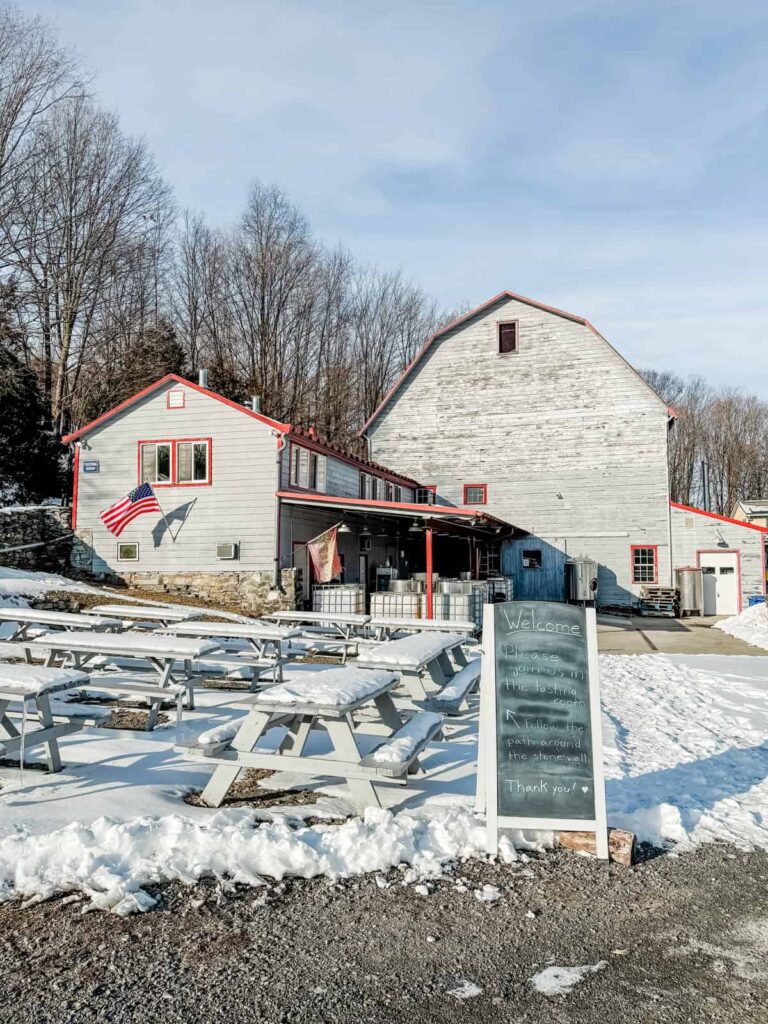 A rustic white barn with red trim at Quartz Rock Vineyard, surrounded by picnic tables covered in snow, with an American flag and a chalkboard sign welcoming guests.