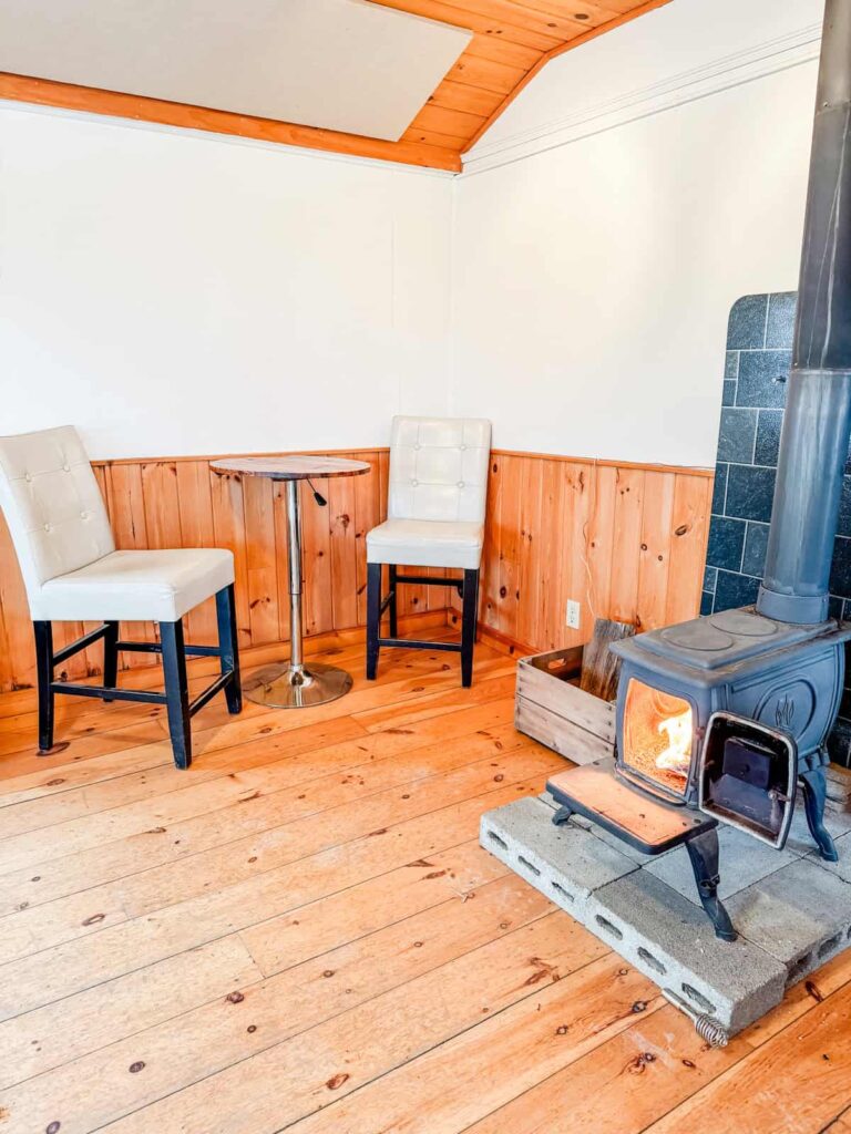 A snug corner inside Quartz Rock Vineyard featuring a small wood-burning stove with a fire inside, a round high-top table, and two cream-colored chairs on wooden floors.
