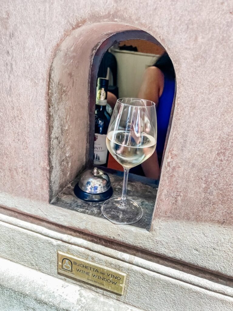 glass of white wine and a service bell rest on the stone ledge of a historic wine window, with a bottle of Chianti partially visible inside the small arched opening.