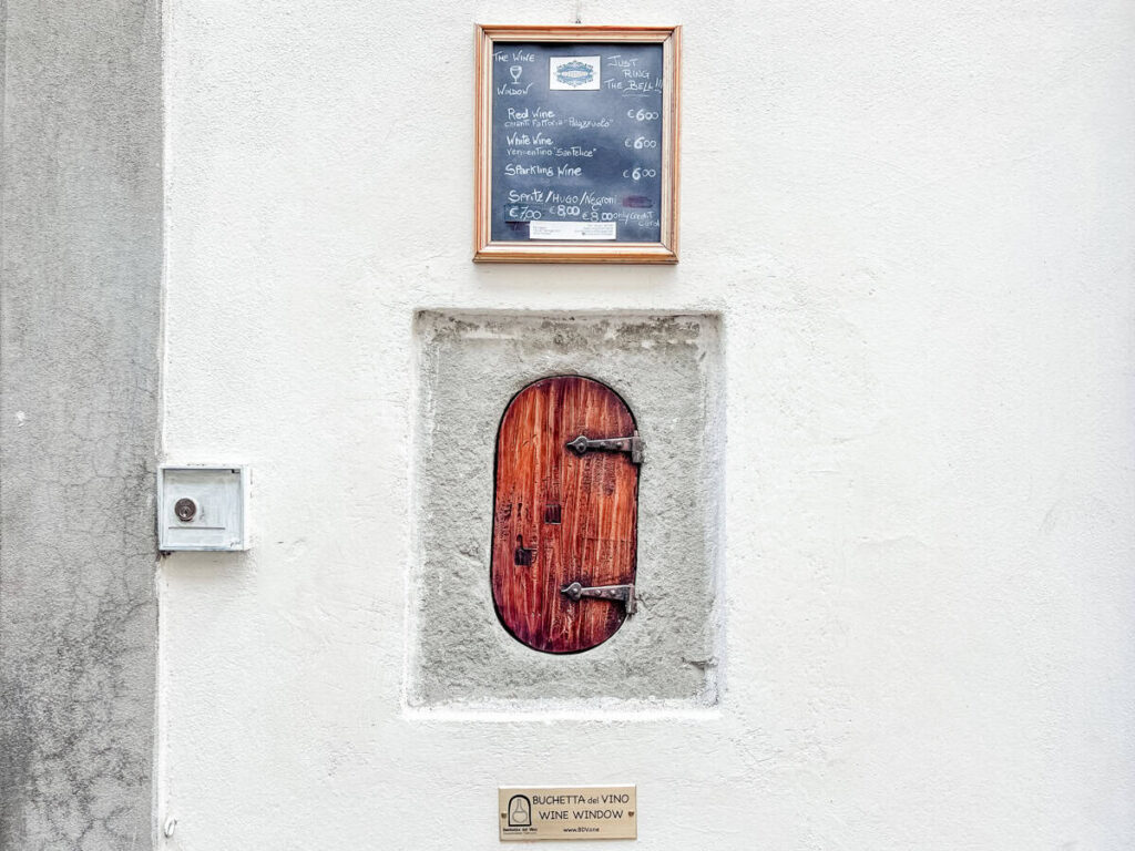 Close-up of Fiaschetteria Fantappiè’s buchetta del vino, a historic wooden wine window with a rustic iron latch, set beneath a framed chalkboard menu listing red, white, and sparkling wines.