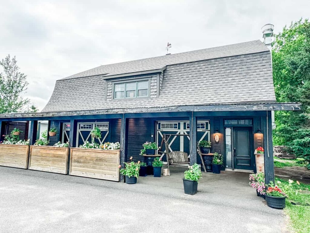 Exterior of Au Pâturage, a rustic restaurant with dark wood siding, a cozy front porch, flower planters, and a pitched roof.