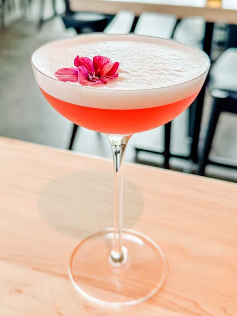 A vibrant pink cocktail at Au Pâturage, topped with a creamy foam and a magenta edible flower, served in a coupe glass on a wooden table.
