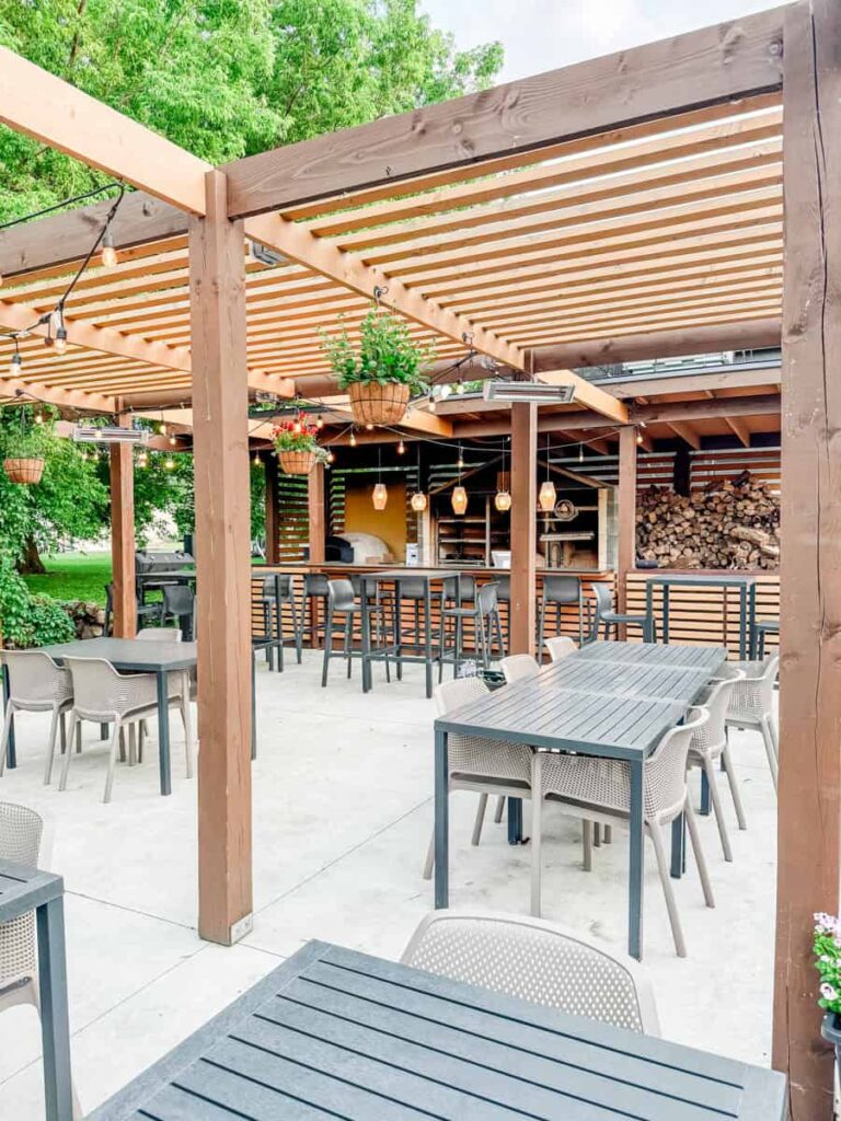 The outdoor patio at Au Pâturage with a modern wooden pergola, string lights, and potted plants above black tables and chairs.
