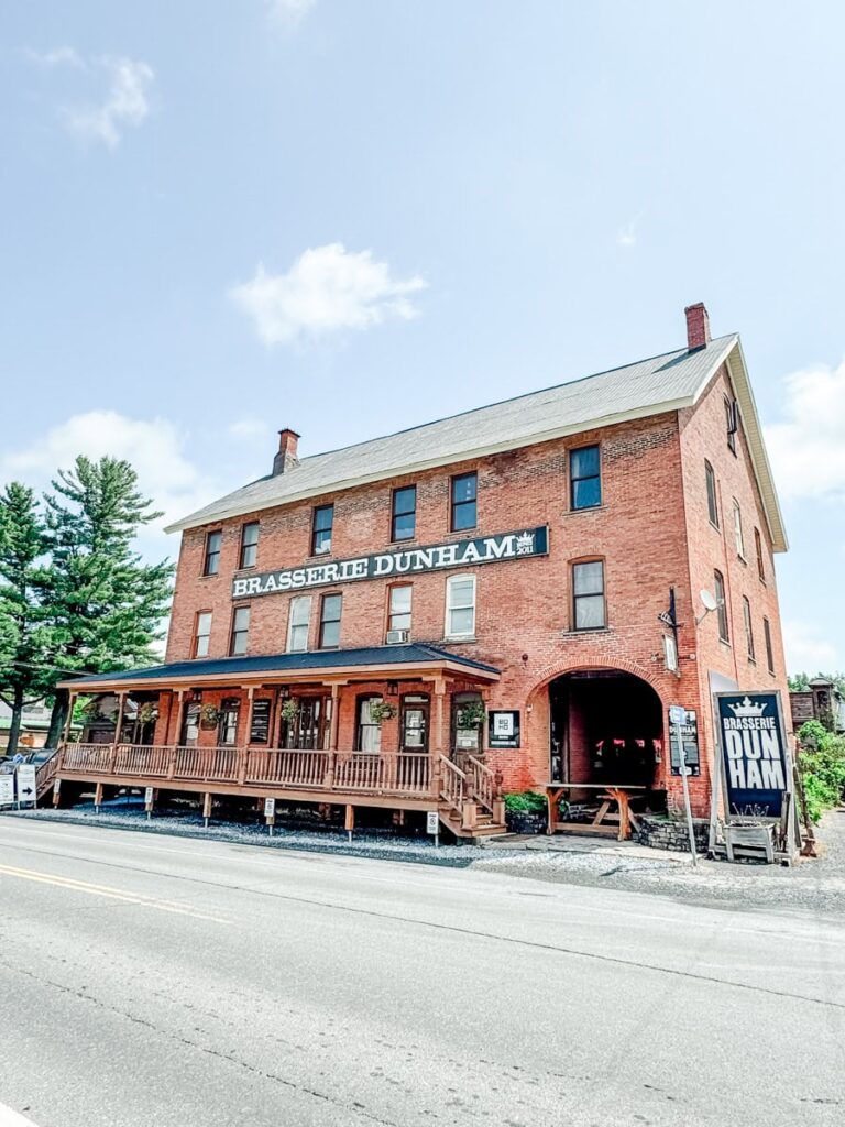 Brasserie Dunham's historic red brick building with a wraparound porch and bold signage, located in the heart of the Eastern Townships.