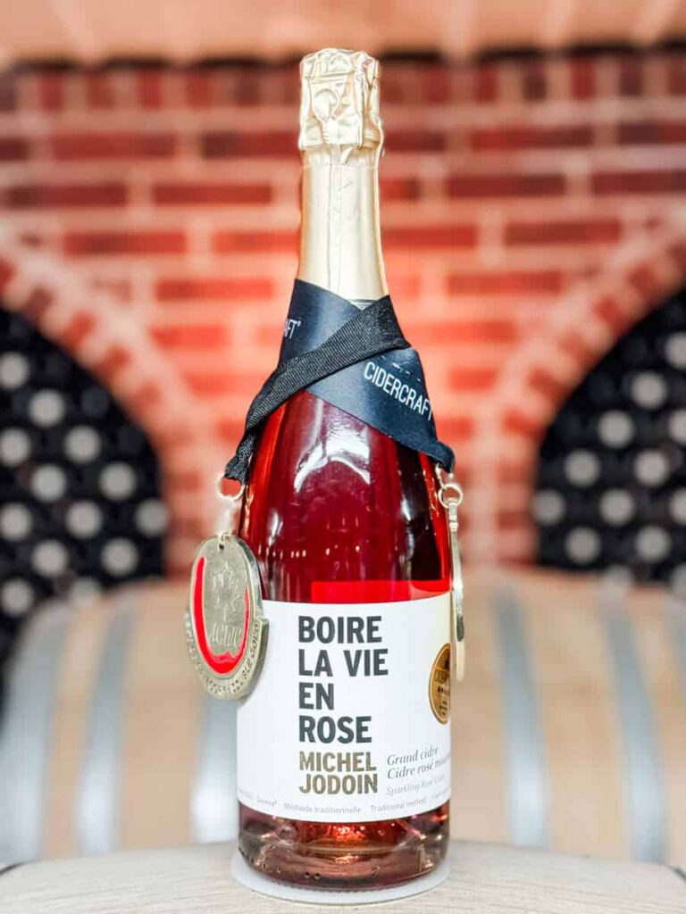 Close-up of a bottle of Michel Jodoin’s sparkling rosé cider, “Boire La Vie en Rose,” displayed on a barrel with two medals around the neck and a brick arch in the background.