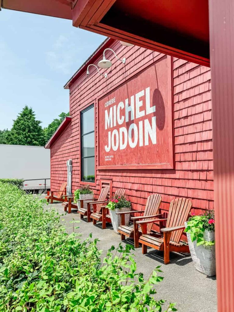 Bright red cider house with large white “Cidrerie Michel Jodoin” lettering on the side, wooden Adirondack chairs out front, and lush greenery lining the walkway.