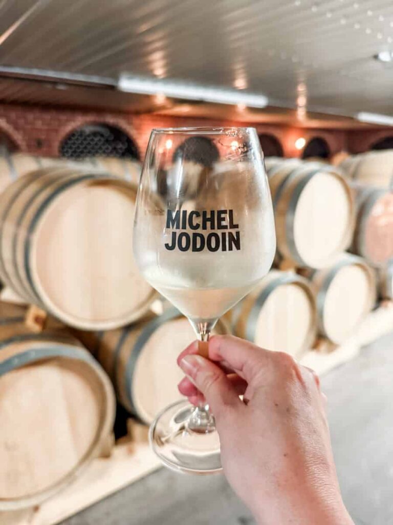 Hand holding a glass of white cider with “Michel Jodoin” printed on it, raised in front of stacked wooden barrels in the cidery’s aging cellar.